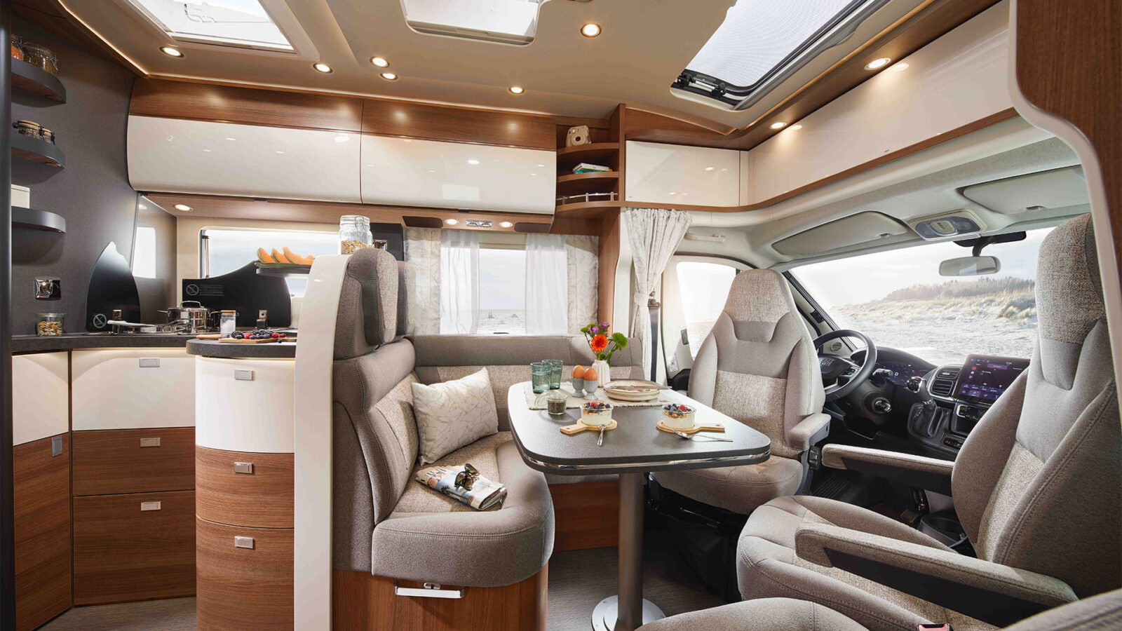 Modernes carthago c1 tourer plus teilintegriertes Wohnmobil-Interieur mit Plüschsitzen, Esstisch mit Lebensmitteln, hellen Deckenleuchten, Holz und weißen Schränken und großen Fenstern, die einen Blick auf den Strand und die Sanddünen im Freien ermöglichen.
