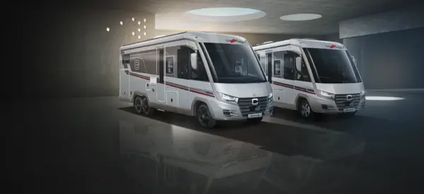 Zwei moderne, silberne Carthago Chic E Line-Wohnmobile mit roten und schwarzen Akzenten stehen im Innenbereich auf einem glänzenden, spiegelnden Boden unter einer mit runden Leuchten und Oberlichtern verzierten Decke.