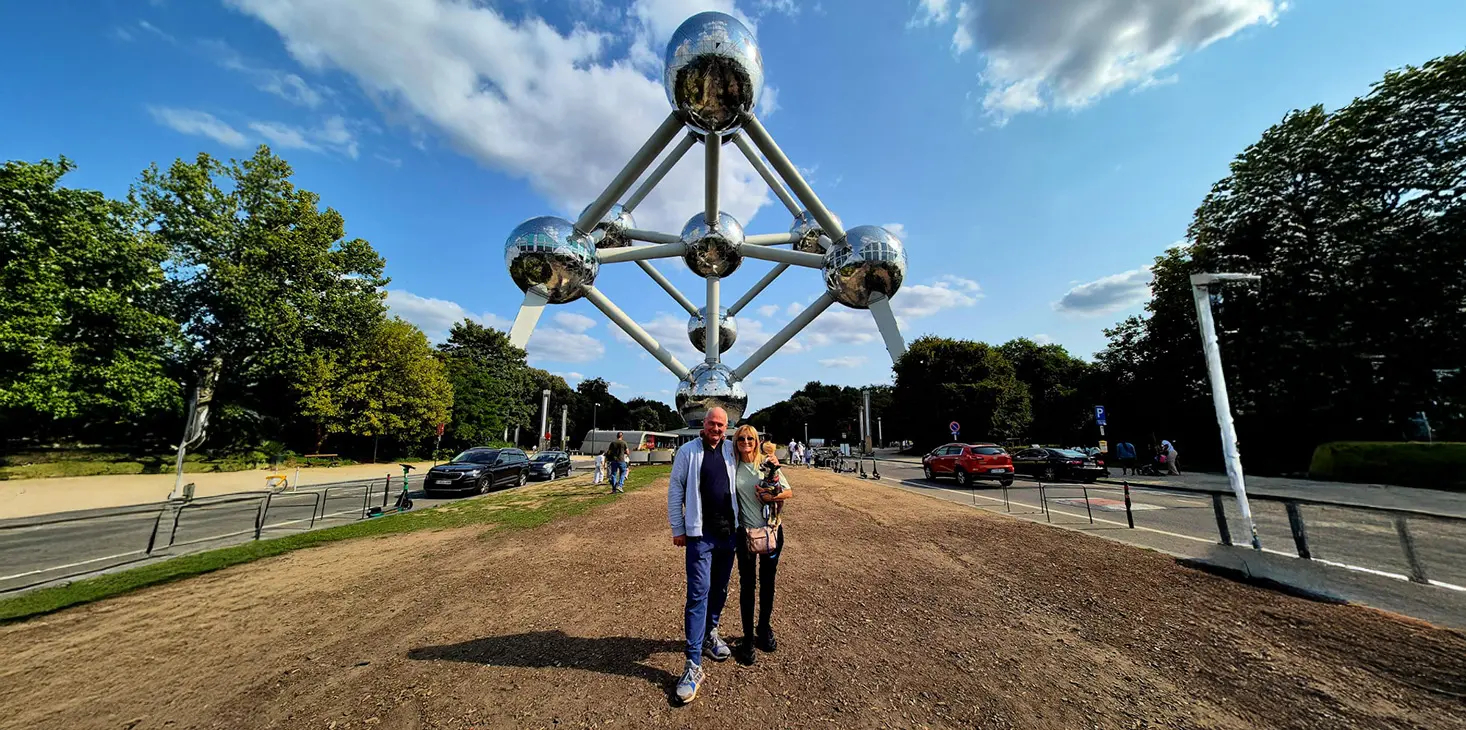 Ein Mann und eine Frau stehen an einem sonnigen Tag mit blauem Himmel und vereinzelten Wolken gemeinsam vor dem Atomium, einer großen Metallstruktur mit verbundenen Kugeln. Im Hintergrund sind Bäume, Autos und Menschen zu sehen.