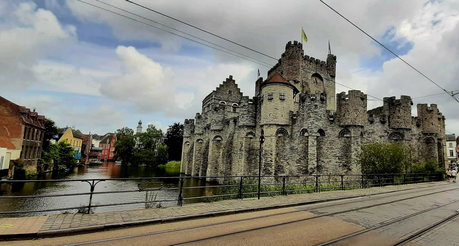 Eine mittelalterliche Steinburg mit runden Türmen und Zinnen steht neben einem Kanal unter einem teilweise bewölkten Himmel, mit Straßenbahnschienen und Gebäuden im Vorder- und Hintergrund.