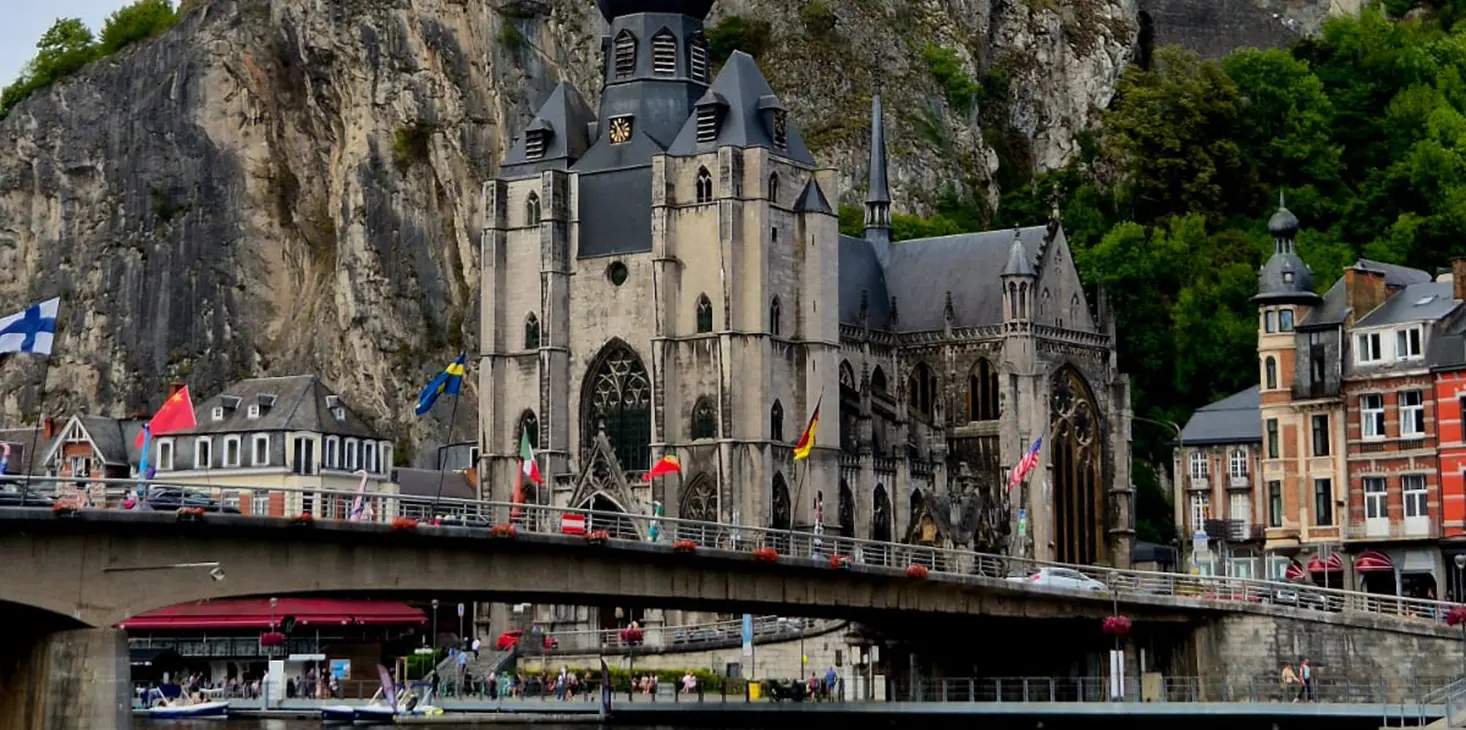 Eine große Kirche im gotischen Stil mit einem hohen Glockenturm steht auf einer Felsklippe, mit einer Brücke und mehreren Fahnen im Vordergrund und bunten Gebäuden an der Seite.