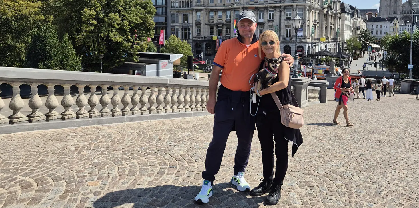 Ein Mann und eine Frau stehen zusammen auf einer Kopfsteinpflasterbrücke und lächeln in die Kamera. Der Mann trägt ein orangefarbenes Hemd und eine Mütze, die Frau trägt Schwarz und eine Sonnenbrille. Im Hintergrund sind städtische Gebäude und Bäume an einem sonnigen Tag zu sehen.