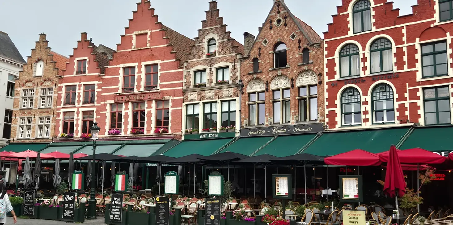 Bunte historische Gebäude mit Treppengiebeln säumen eine Straße in Brügge, Belgien. Darunter befinden sich Restaurants mit grünen und roten Markisen, die mit Sitzplätzen im Freien und Menütafeln eine lebendige, einladende Atmosphäre schaffen.