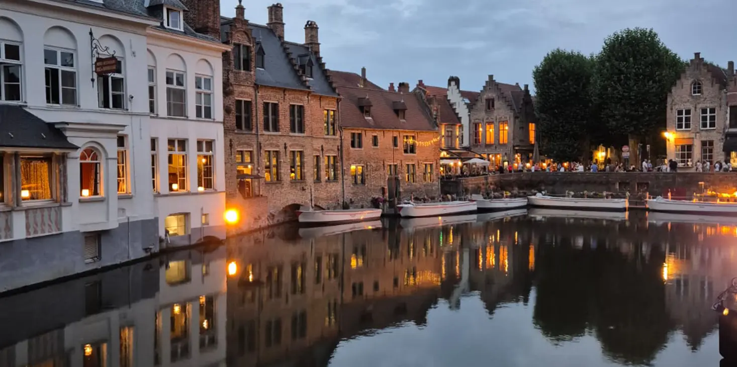 Ein von historischen Backsteingebäuden gesäumter Kanal, der in der Abenddämmerung warm beleuchtet ist und dessen Lichter sich im ruhigen Wasser spiegeln. Auf der rechten Seite sind Sitzgelegenheiten im Freien und Bäume zu sehen, die ein friedliches europäisches Stadtbild schaffen.