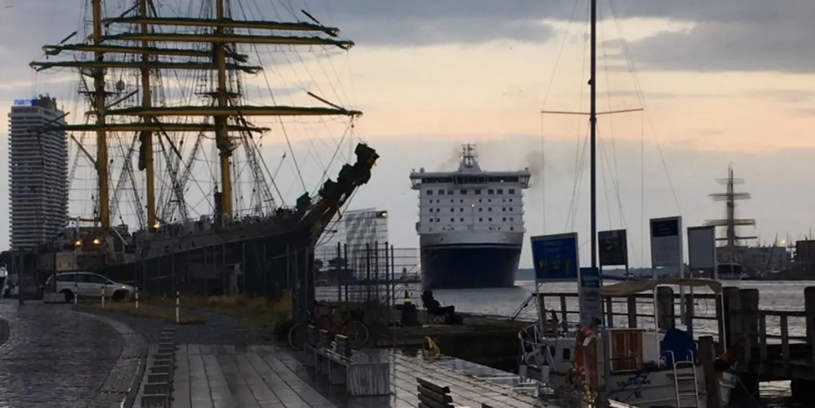 Ein großes Kreuzfahrtschiff nähert sich in der Abenddämmerung einem Hafen. Links ist ein großes Segelschiff angedockt, im Hintergrund sind moderne Gebäude zu sehen, und der Himmel ist bewölkt.