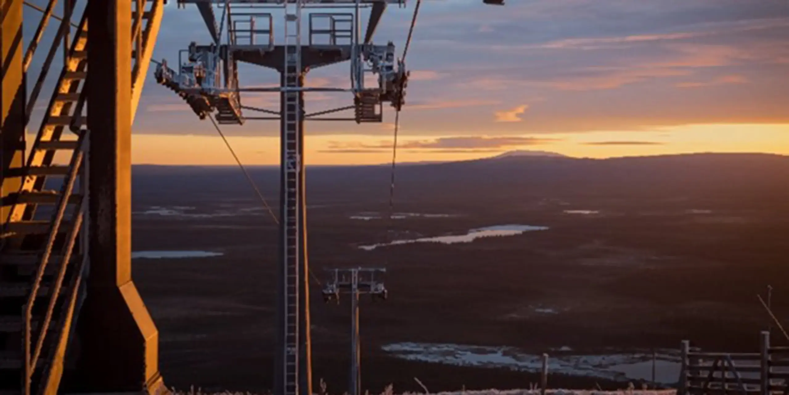 Ein Skiliftturm steht bei Sonnenuntergang auf einem Berg und überblickt eine weite Landschaft mit Flüssen, Wäldern und fernen Hügeln unter einem bunten Himmel.