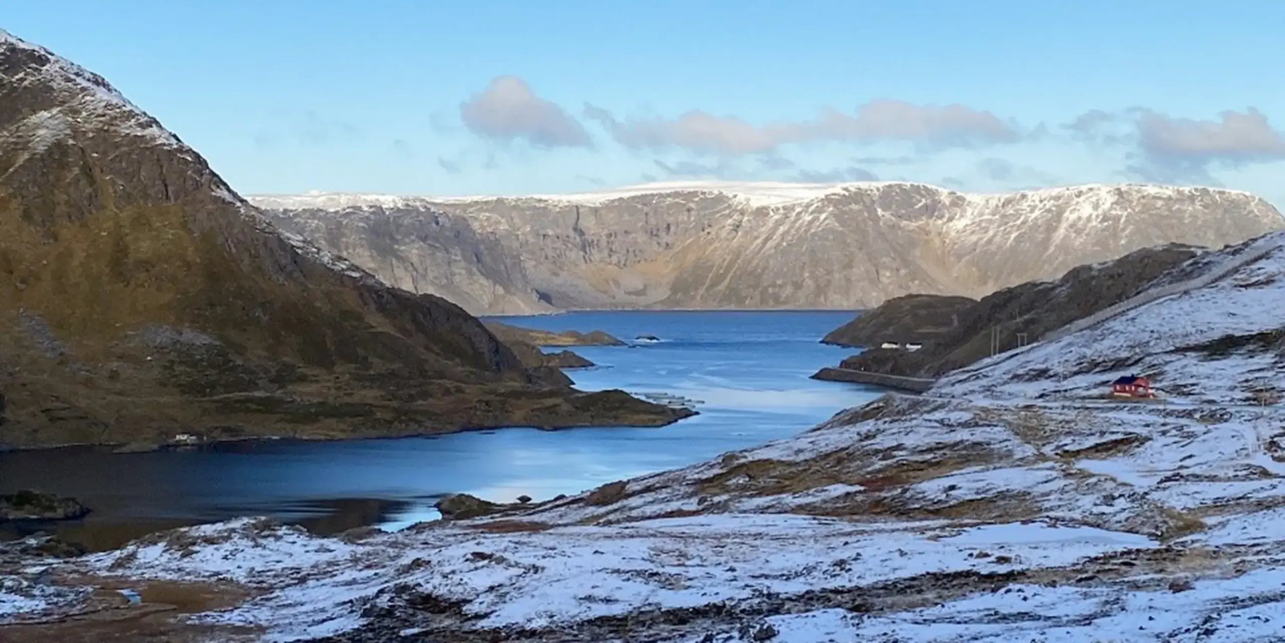 Ein malerischer Fjord mit ruhigem, blauem Wasser, umgeben von schneebedeckten Bergen unter einem klaren Himmel, mit einem kleinen roten Haus am rechten Hang.