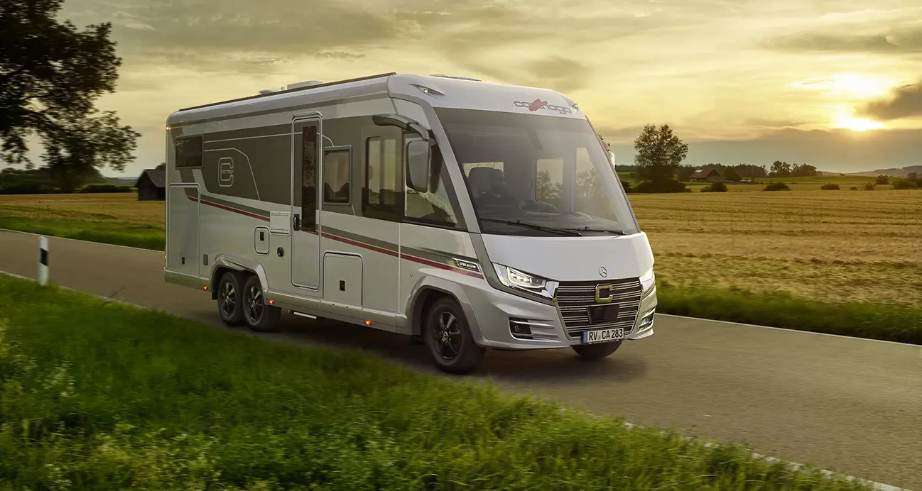 Ein modernes silbernes Wohnmobil, frisch nach der Gasprüfung im Wohnmobil, fährt bei Sonnenuntergang auf einer ländlichen Straße entlang, vorbei an Feldern und Bäumen unter einem teils bewölkten Himmel.
