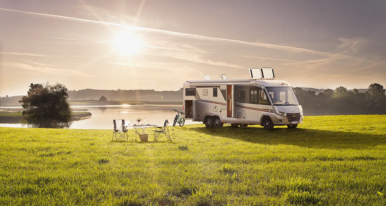 Ein Wohnmobil ist auf einer Wiese in der Nähe eines ruhigen Flusses bei Sonnenuntergang geparkt. Zwei Stühle, ein Tisch und ein Fahrrad sind draußen aufgestellt, um eine friedliche Campingszene zu schaffen, mit Bäumen und sanften Hügeln im Hintergrund - perfekt für Ihre Gasprüfung im Wohnmobil.