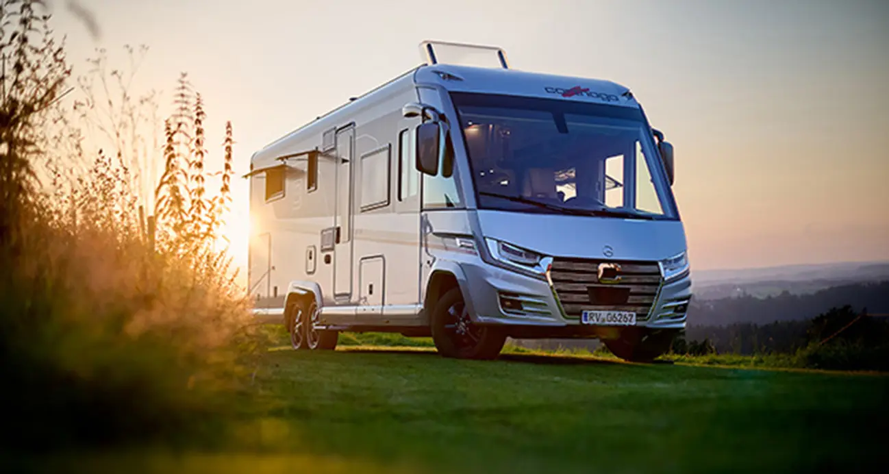 Ein weißes Wohnmobil, perfekt für Langstrecken im Wohnmobil, ist bei Sonnenuntergang auf einem grasbewachsenen Hügel geparkt. Das Sonnenlicht strömt durch seine Fenster und beleuchtet das hohe Gras im Vordergrund unter einem klaren, goldenen Himmel.