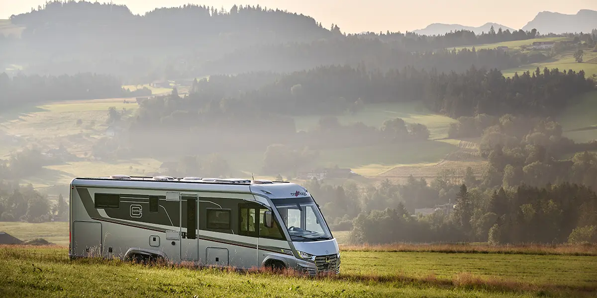 Ein modernes Wohnmobil, perfekt für Langstrecken im Wohnmobil, steht auf einer grasbewachsenen Wiese mit sanften Hügeln, Wäldern und fernen Bergen im Hintergrund unter einem dunstigen Himmel.