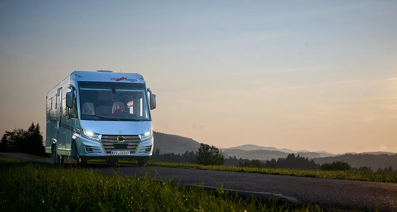 Ein weißes Wohnmobil fährt bei Sonnenuntergang eine Landstraße entlang, perfekt für Langstrecken im Wohnmobil, mit grünem Gras im Vordergrund und sanften Hügeln und Bäumen in der Ferne unter einem klaren Himmel.