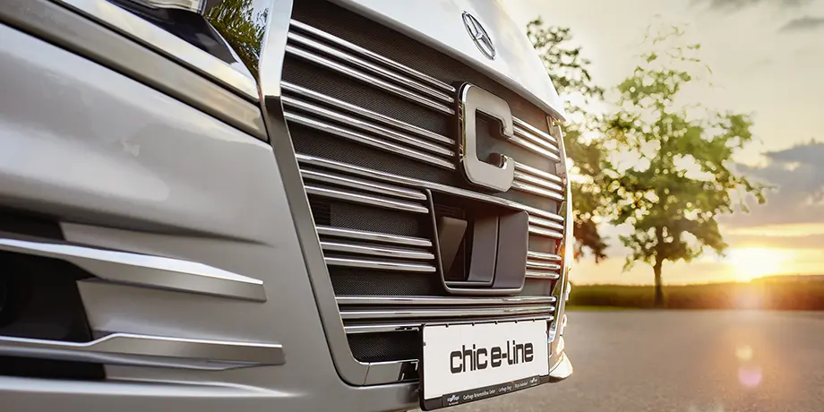 Nahaufnahme des vorderen Kühlergrills und der Stoßstange eines silbernen Fahrzeugs mit einem Nummernschild mit der Aufschrift chice-line vor einem Sonnenuntergang und einem verschwommenen Baum - perfekt für die Vorbereitung auf die Gasprüfung im Wohnmobil-Abenteuer.