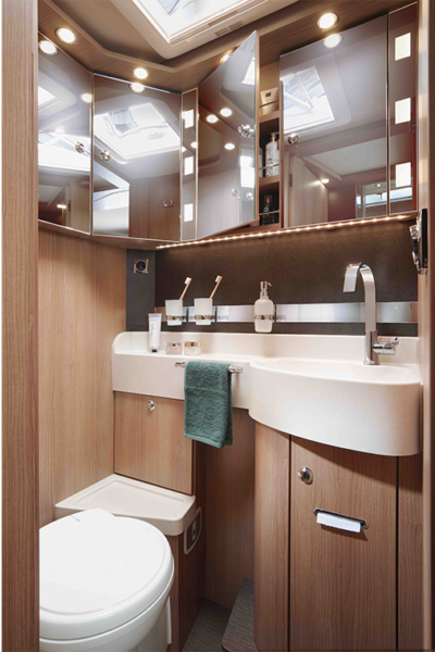 Modernes kompaktes Bad im Carthago C Tourer mit Holzverkleidung, weißem Waschbecken, Spiegelschränken, Toilette, wandmontierten Toilettenartikeln und einem grünen Handtuch auf dem Waschbecken. Einbaulampen und Oberlichter sorgen für helle Beleuchtung.