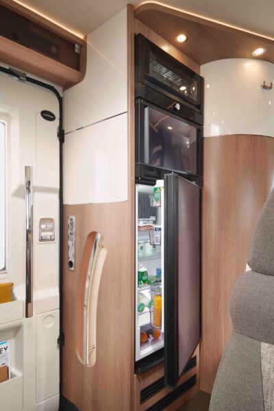 Ein modernes Carthago C1-tourer Edition+ T Teilintegriertes Wohnmobil mit eingebautem Kühl- und Gefrierschrank. Beide Türen lassen sich öffnen und geben den Blick auf Regale mit Lebensmitteln und Getränken frei. Helle Holzverkleidungen, ein grauer Sitz und ein Teil der Eingangstür sind ebenfalls zu sehen.