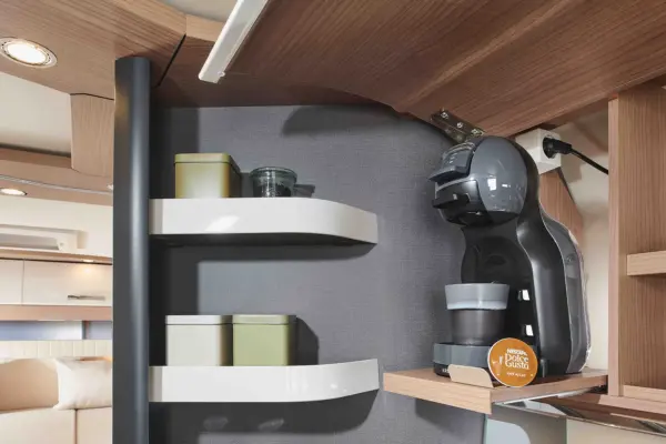 Eine moderne Küchenecke in einem Carthago C Tourer zeigt Wandregale mit Behältern und eine Kapselkaffeemaschine, die ein Getränk aufbrüht, eine Dolce Gusto-Pad, die inmitten von hellem Holz und grauem Dekor zu sehen ist.