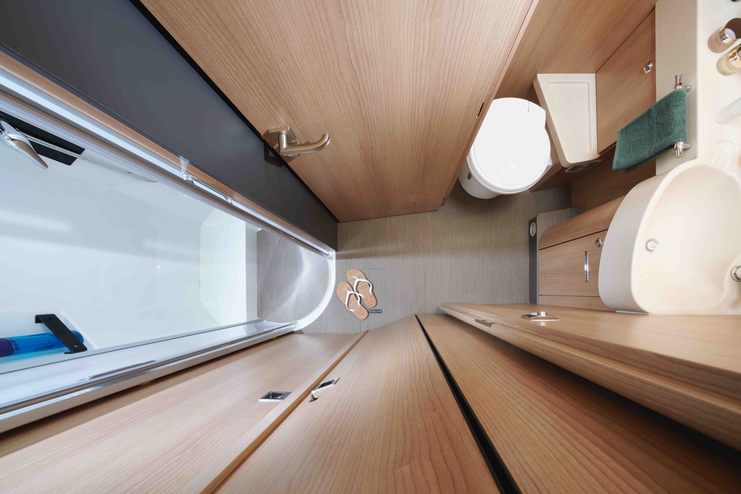 Vue aérienne d'une salle de bains compacte avec des surfaces en bois, des toilettes blanches, un lavabo, une serviette verte, des pantoufles sur le sol et une douche avec une porte en verre. L'espace semble bien organisé et propre.