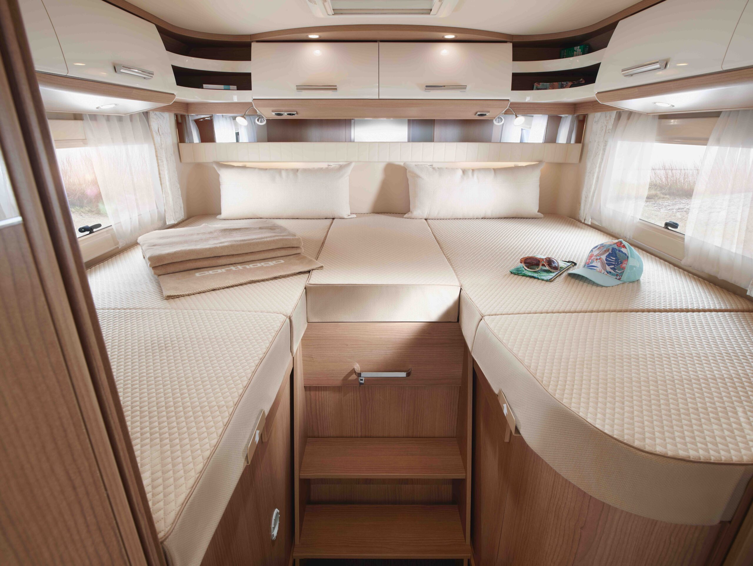 Intérieur d'un camping-car où l'on trouve un lit bien fait avec une literie beige, des serviettes pliées, un chapeau et une assiette de snacks sur le dessus ; des armoires en bois, des étagères et des rideaux complètent l'espace confortable et compact.