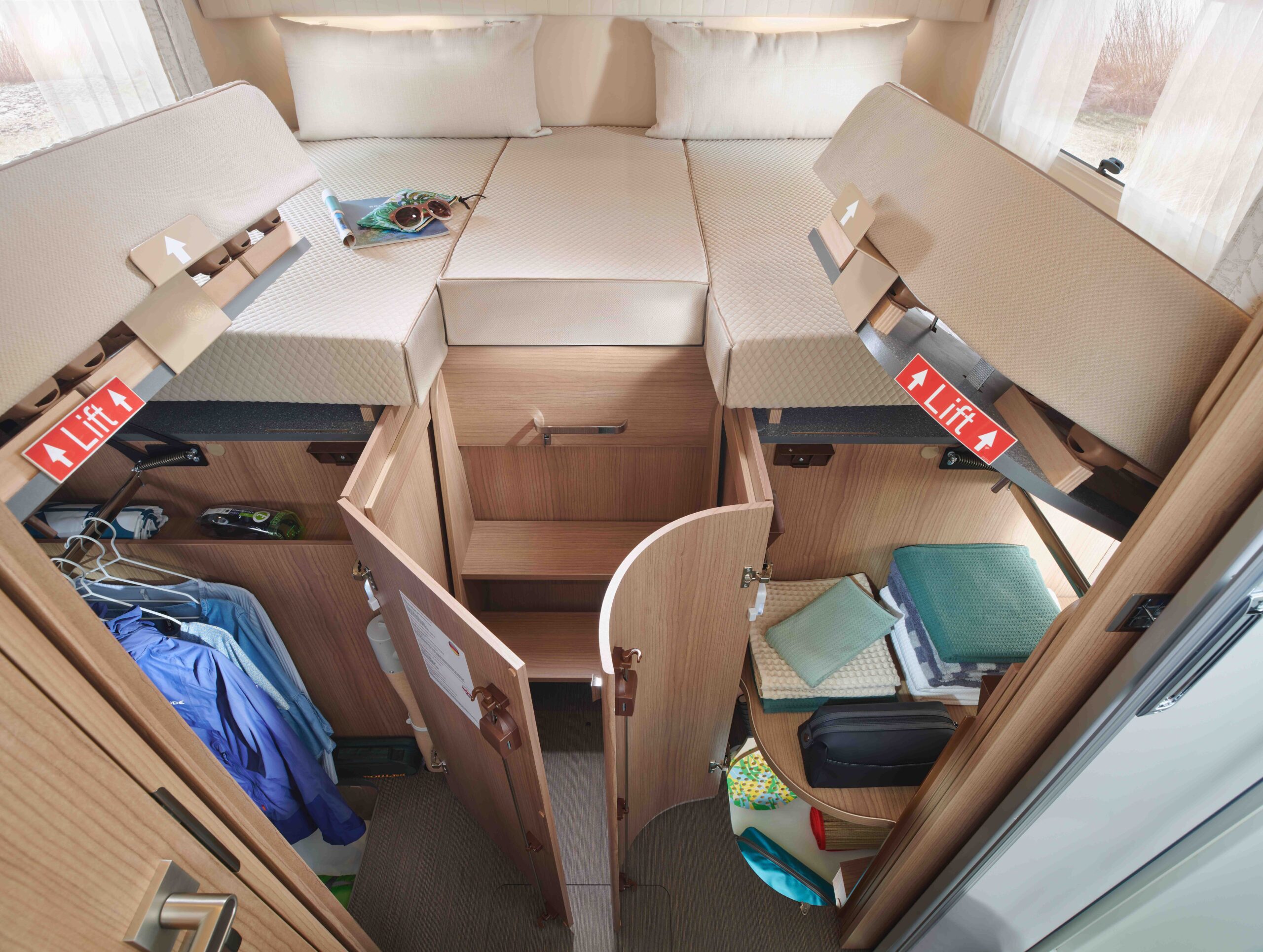 Intérieur d'un camping-car montrant un espace de rangement organisé sous deux lits jumeaux surélevés, avec des vêtements, des livres, des appareils électroniques et d'autres objets personnels soigneusement rangés dans des compartiments et des armoires.
