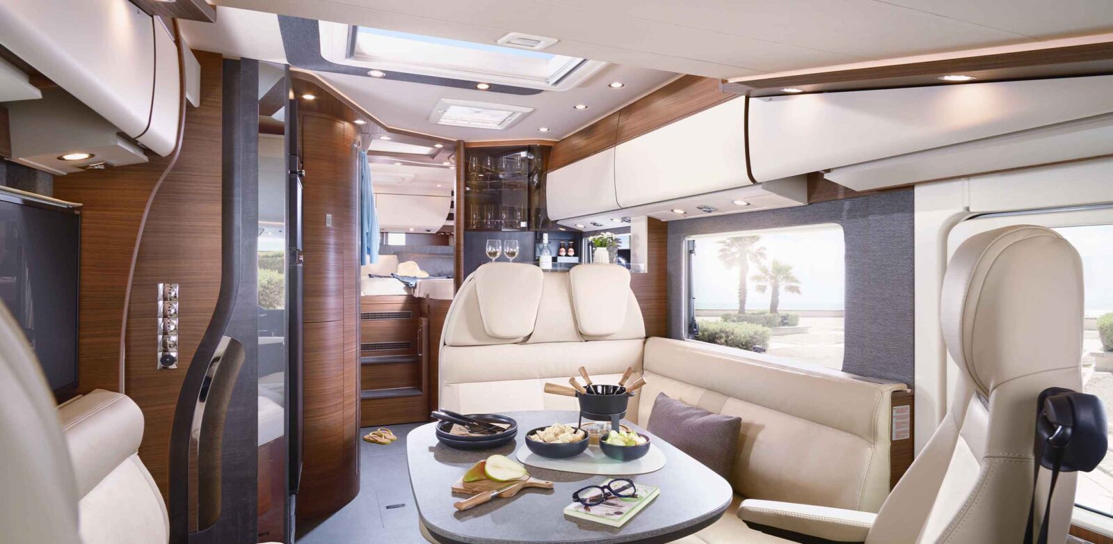 Modernes Wohnmobil-Interieur mit cremefarbenen Ledersitzen, einem Esstisch mit Speisen und Getränken und Holzverkleidung. Große Fenster geben den Blick auf einen sonnigen Strand mit Palmen frei - und unterstreichen das Neuheiten-Modelljahr in stilvollem Komfort.