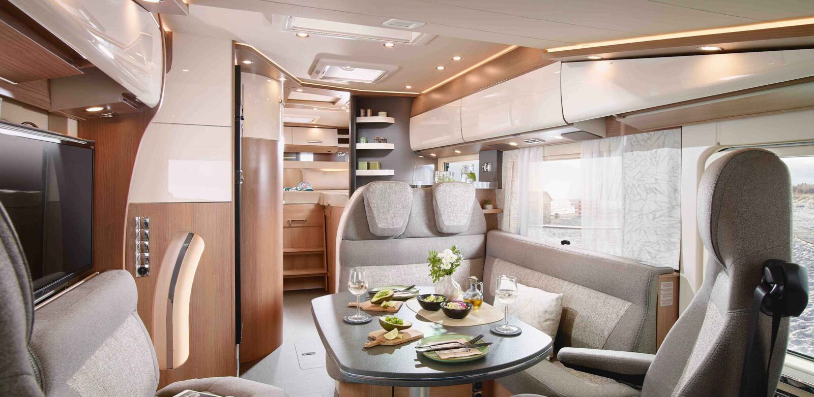 Ein modernes, helles Wohnmobil-Interieur aus dem Neuheiten Modelljahr mit einem gedeckten Esstisch mit Speisen und Getränken, grauen gepolsterten Sitzen, Stauraum über Kopf und einem kompakten Küchenbereich im Hintergrund.