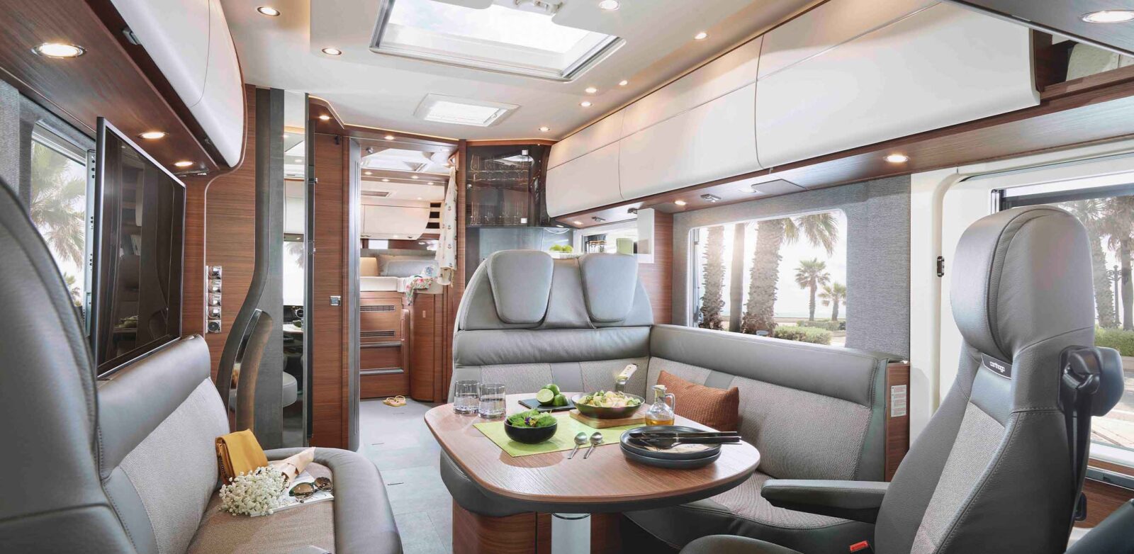 Modernes Wohnmobil-Interieur im Neuheiten Modelljahr, mit einem Essbereich für zwei Personen, gepolsterten Sitzen, einem Tisch mit Salat und Getränken, Stauraum über dem Kopf und großen Fenstern, die Palmen und Sonnenlicht nach draußen lassen.