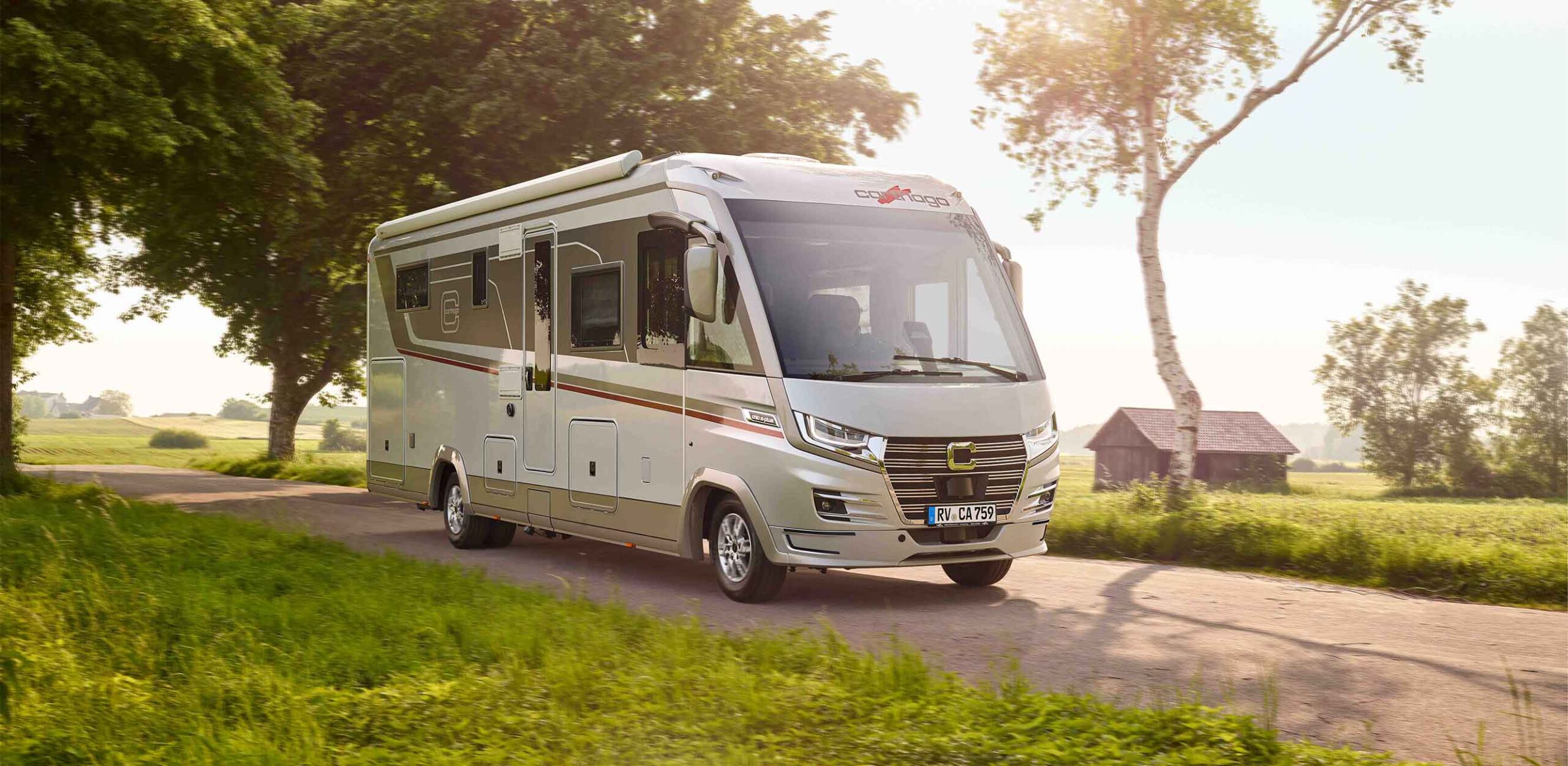 Ein großes, modernes weißes Wohnmobil mit Partnervorstellung Truma fährt auf einer Landstraße, umgeben von grünem Gras, Bäumen und einer Holzscheune, unter strahlendem Sonnenschein.
