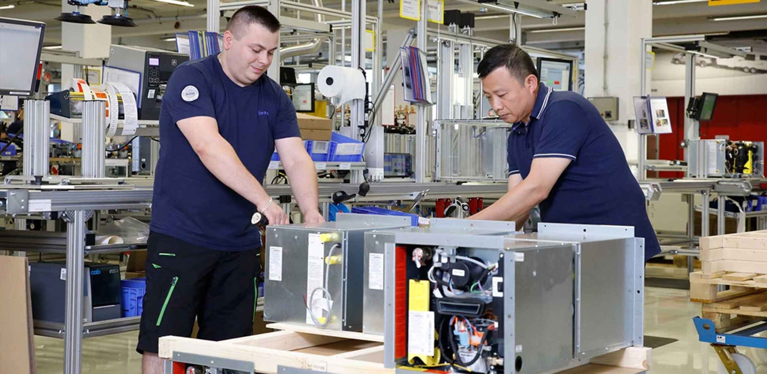 Zwei Männer montieren oder prüfen in einer Fabrikumgebung große elektrische Metallteile auf einer Werkbank, umgeben von Industrieanlagen und Werkzeugen, während einer Veranstaltung der Partnervorstellung Truma.