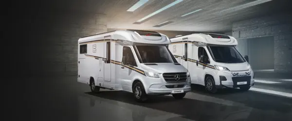 Zwei moderne weiße Wohnmobile mit schlichtem Design stehen nebeneinander in einem geräumigen, gut beleuchteten Innenraum mit grauen Wänden und polierten Böden.