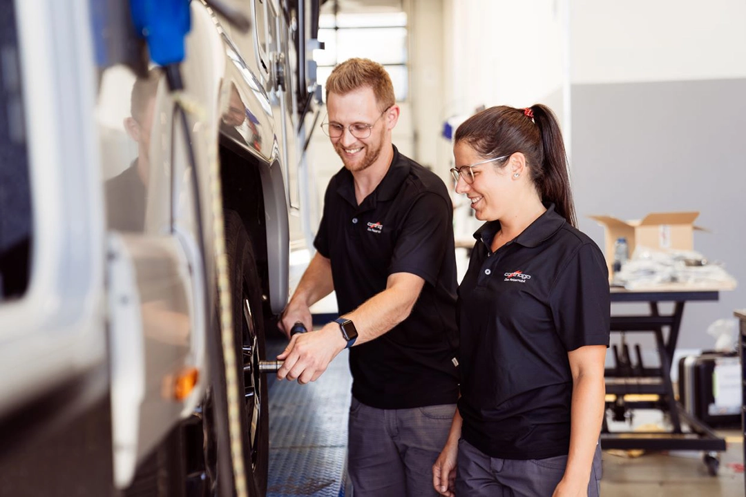 Zwei Personen in passenden schwarzen Hemden arbeiten gemeinsam an einem großen Autoreifen in einer gut beleuchteten Garage oder Werkstatt. Eine Person bearbeitet den Reifen mit einem Werkzeug, während die andere lächelnd zusieht.