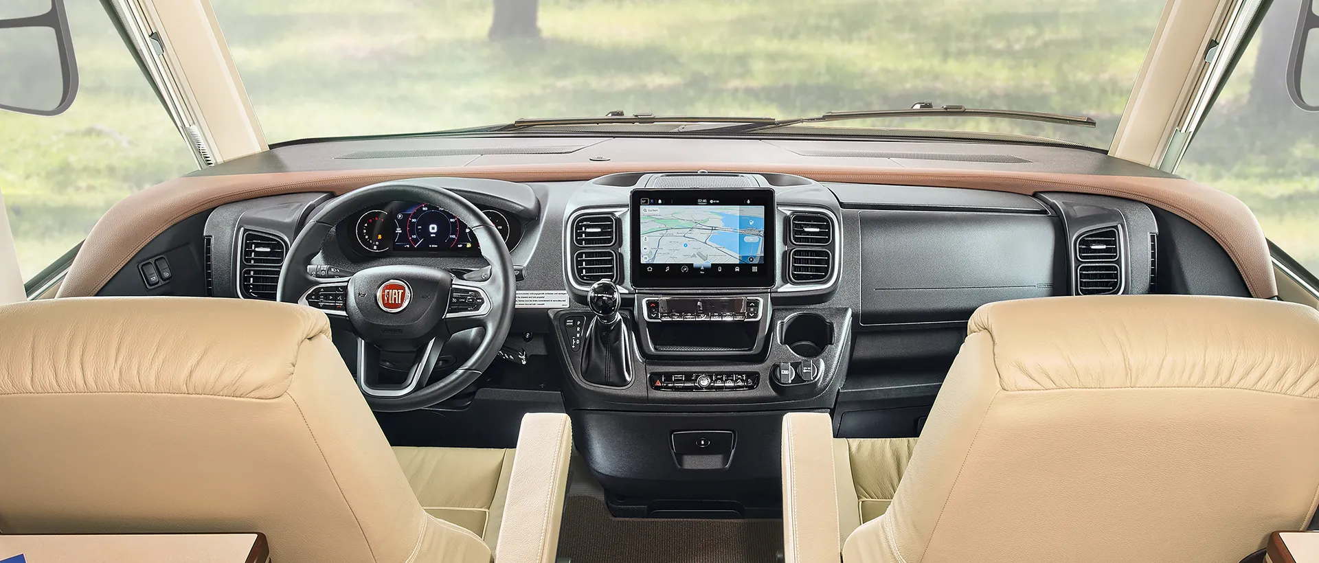 Blick von zwei beigen Kapitänssesseln auf das Armaturenbrett eines Fiat-Reisemobils, das auf einem robusten Fahrgestell für Wohnmobile aufgebaut ist, mit Touchscreen-Navigationssystem, Lenkrad und Bedienelementen, mit einer großen Windschutzscheibe, durch die man ins Grüne schauen kann.