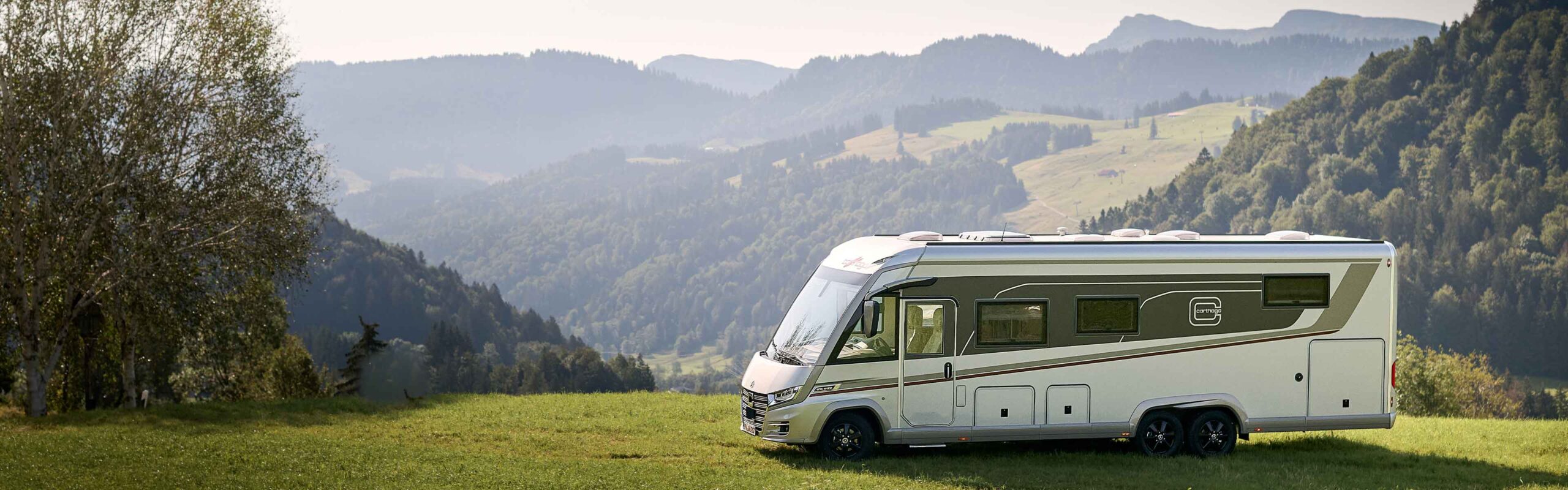 Ein großes weißes und graues Wohnmobil steht auf einem grasbewachsenen Feld mit sanften Hügeln und bewaldeten Bergen im Hintergrund unter einem klaren Himmel.