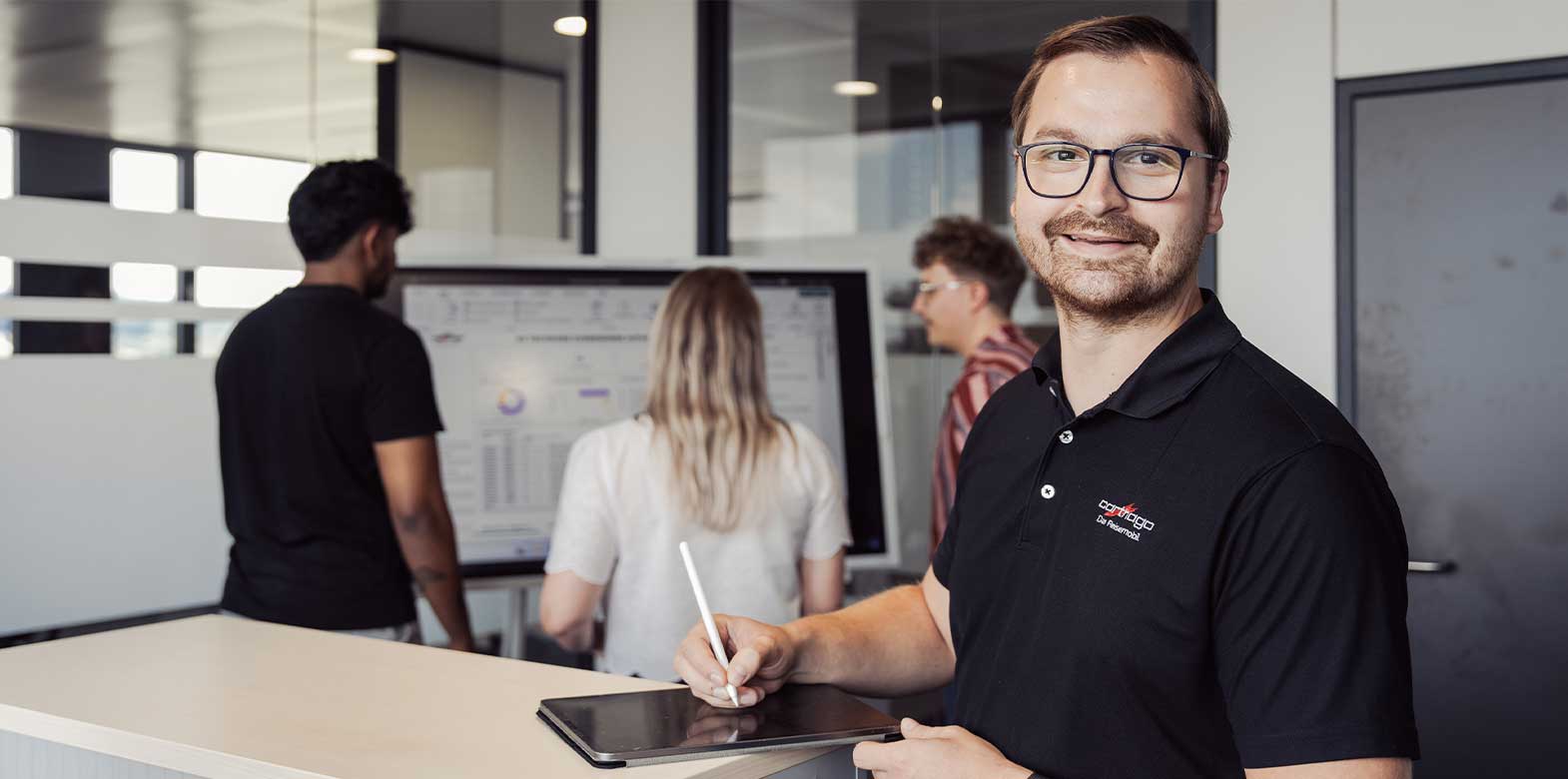 Ein Mann mit Brille lächelt in die Kamera, während er einen Stift und ein Tablet in der Hand hält; hinter ihm stehen drei Kollegen vor einem Bildschirm, auf dem Diagramme in einer modernen Büroumgebung angezeigt werden.