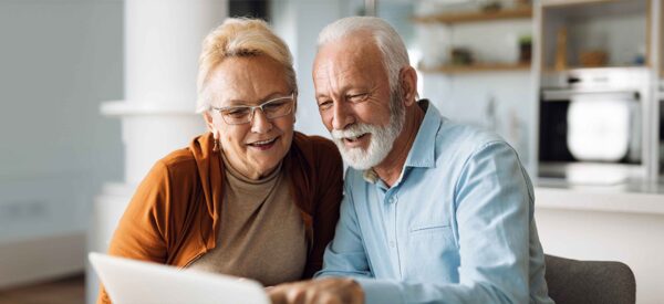 An elderly couple sits close together at a table, smiling as they look at a laptop screen in a bright, modern kitchen.