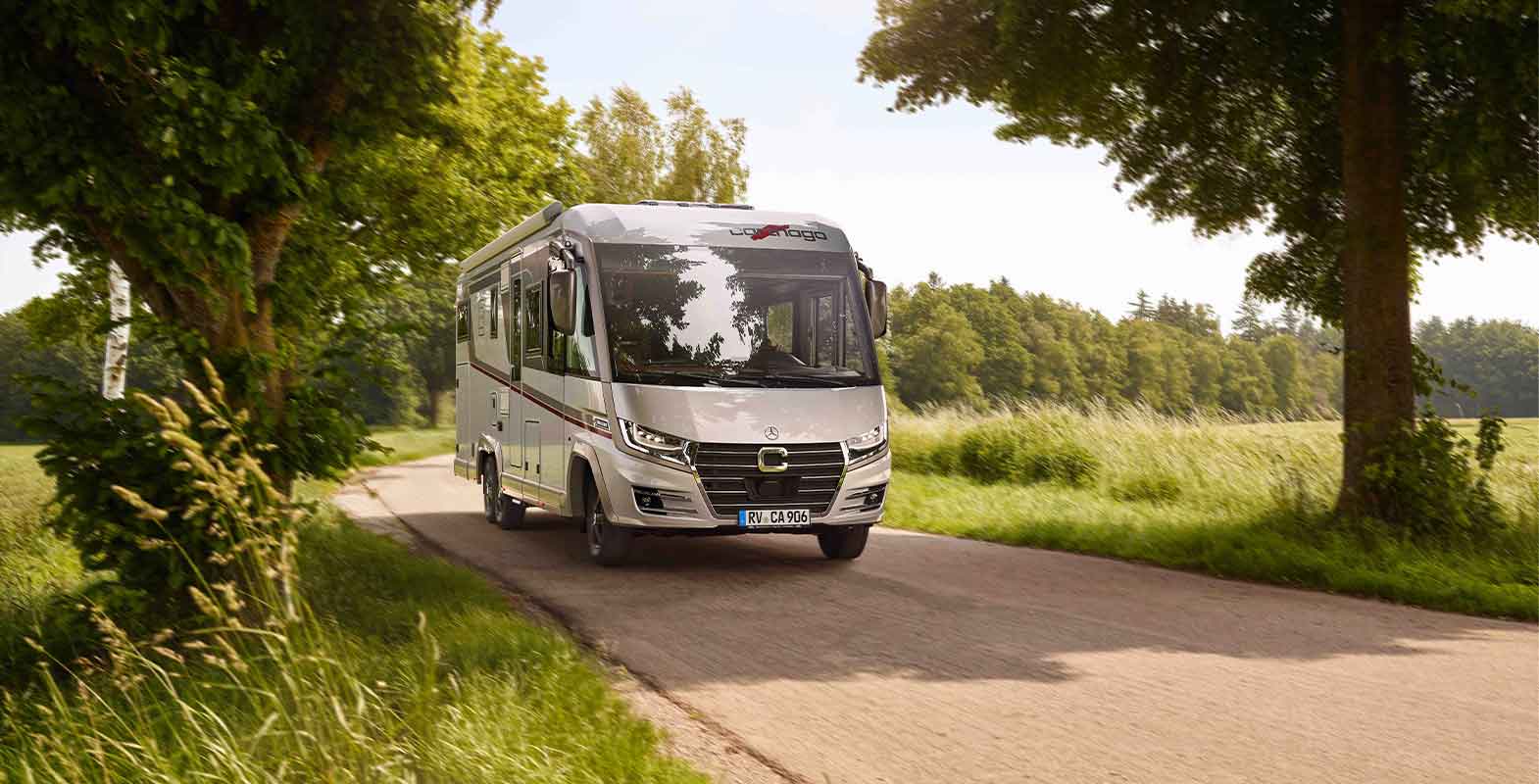 Ein silbernes Wohnmobil fährt auf einer schmalen, von Bäumen gesäumten Landstraße mit grasbewachsenen Feldern und üppig grünen Bäumen auf beiden Seiten unter einem teilweise sonnigen Himmel.