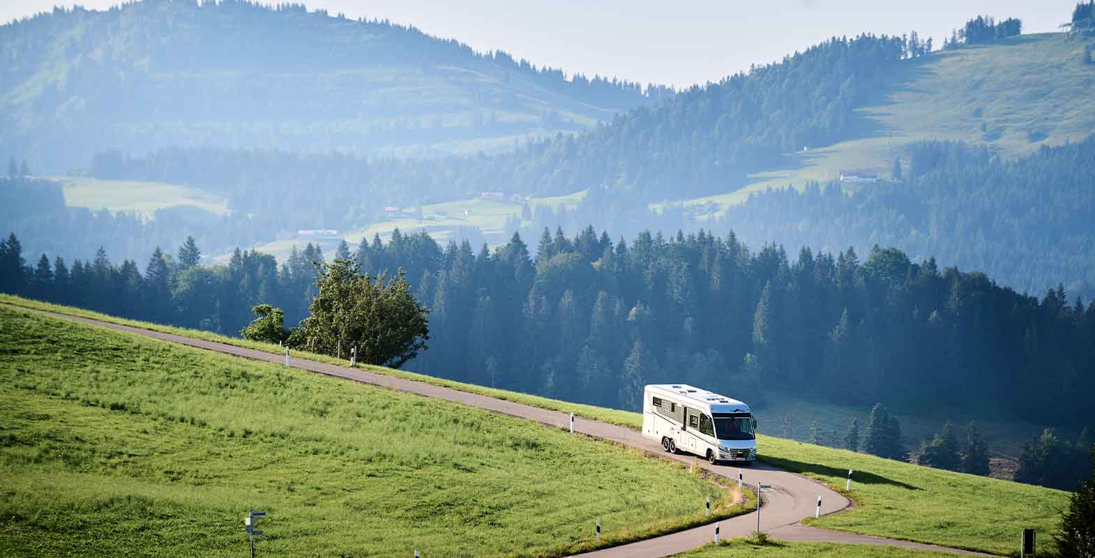 Ein weißes Wohnmobil fährt auf einer kurvenreichen Straße durch grüne Hügel, umgeben von Bäumen und einer vielschichtigen Berglandschaft unter einem dunstigen Himmel.