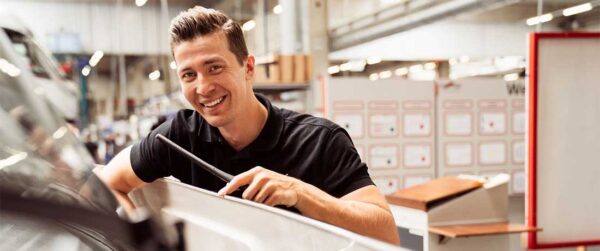 Ein lächelnder Mann in einem schwarzen Hemd arbeitet in einer hell erleuchteten Autowerkstatt an einem Autoscheibenwischer.