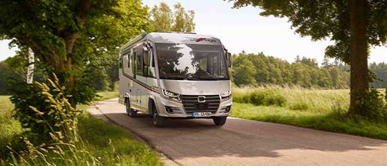 Ein silbernes Wohnmobil fährt eine schmale Landstraße entlang, umgeben von grünem Gras, Bäumen und hellem Tageslicht.