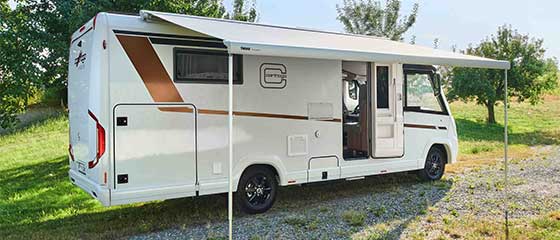 Ein weißes Wohnmobil mit braunen und schwarzen Akzenten ist auf Gras und Schotter geparkt. Die seitliche Markise ist ausgefahren und spendet Schatten, und im Hintergrund sind Bäume zu sehen.