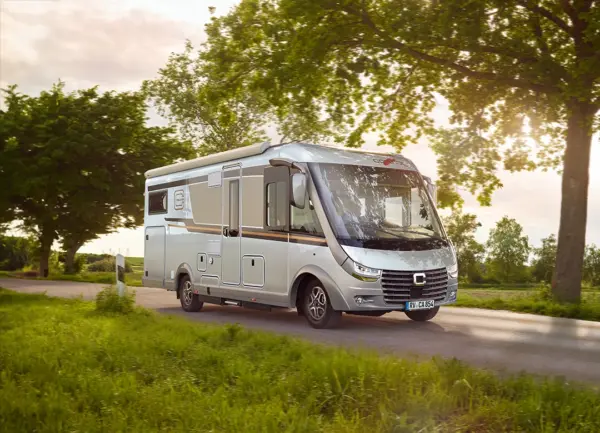 A white motorhome is driving along a country road lined with green grass and trees, with sunlight filtering through the leaves and a clear sky in the background.